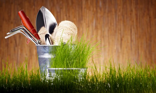 A close-up image of a small garden scene set on a wooden surface, featuring a metal container filled with gardening tools and supplies. Inside the container, there is a pair of hedge shears with red handles, a hand trowel, a gardening fork, and a roll of natural twine. Surrounding the container, there are vibrant green grass blades, creating a lush, natural backdrop that highlights outdoor gardening activity. The wooden background provides a warm, rustic tone, enhancing the natural feel of the scene. This setup reflects typical gardening supplies used for landscaping and maintenance work at residential gardens or outdoor spaces near Chelsea, emphasizing the importance of basic tools in professional gardening services such as those offered by Gardening Services Chelsea. The lighting suggests a bright, possibly sunny day, ideal for outdoor gardening and lawn care. The image underscores essential gardening elements and tools relevant to lawn and garden maintenance, supporting local SEO for gardening services in Chelsea and the surrounding area.